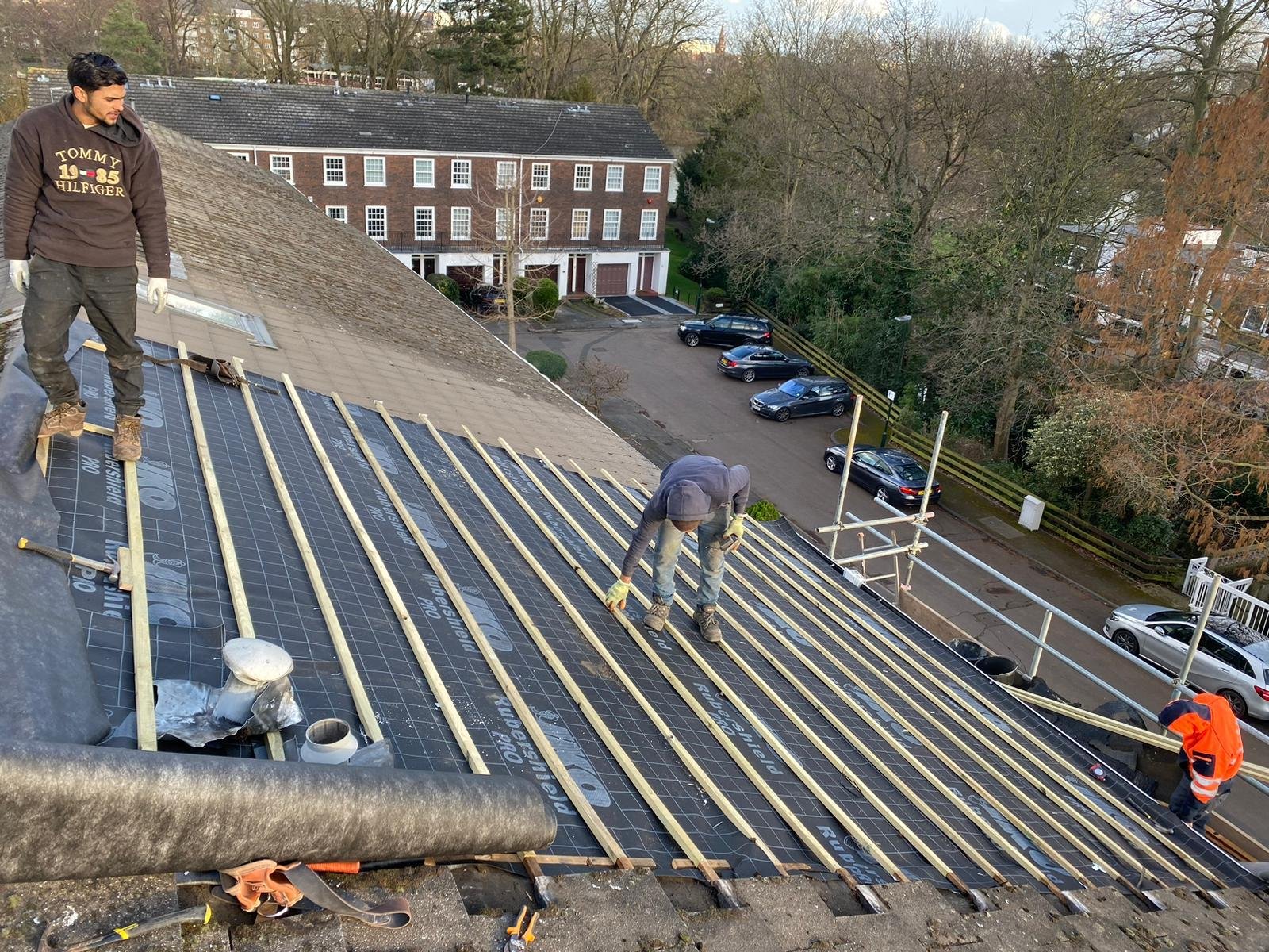 Workers installing solar panels on a residential roof, with a brick building and parked cars visible in the background
