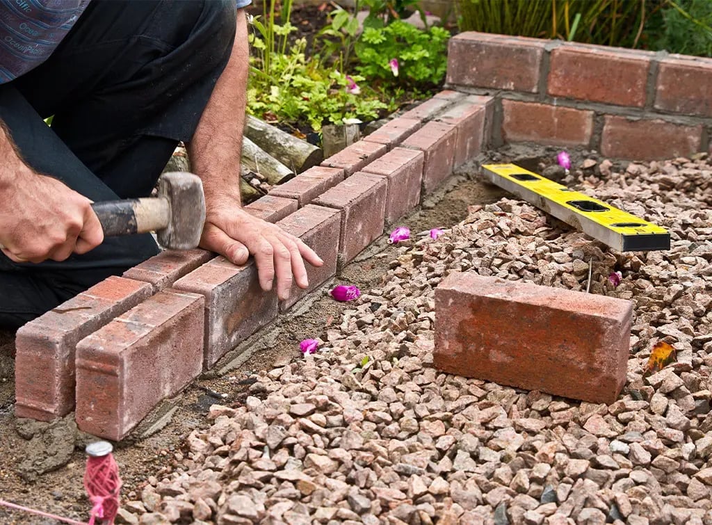 Person laying bricks on gravel in a garden bed with purple flowers and green foliage visible