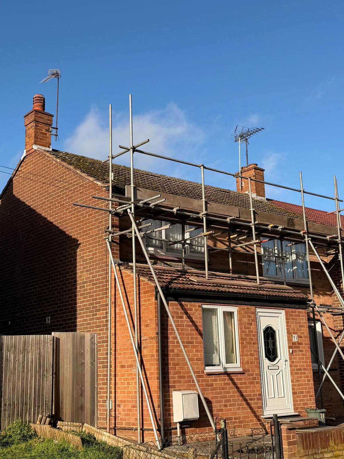 Red brick house under construction with metal scaffolding covering the roof and upper section against a blue sky