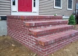 Red brick front porch steps leading to a house with gray siding and a magenta door