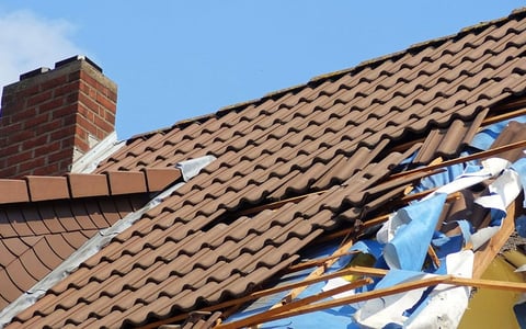 Damaged roof with torn tiles and exposed underlayment, blue tarping covering holes